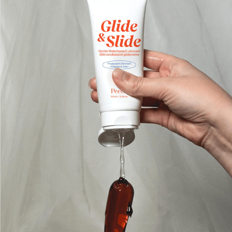 En hand klämmer ut vattenbaserat Glide & Slide glidmedel från tuben på en mörk sexleksak