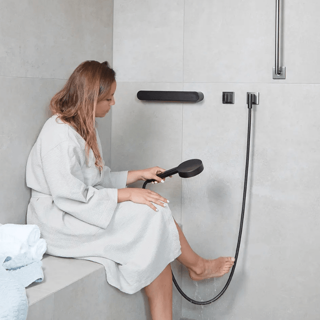 Kvinde i badekåbe sidder på en bænk i badet med et sort støttegreb monteret på væggen