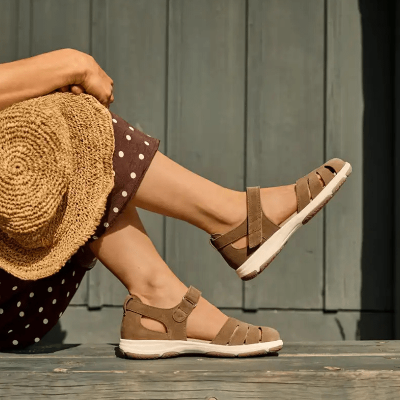 En kvinna med bruna New Feet damsandaler i nubuck på fötterna, sittande på en träbänk med en stråhatt