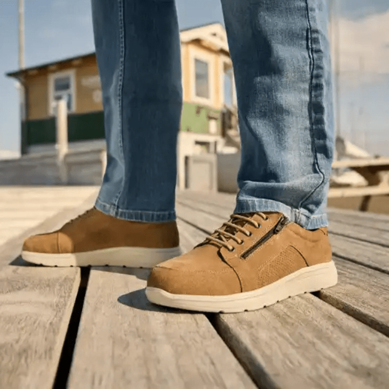 En person i blå jeans som bär New Feet herrskor i mocka, stående på en träbrygga utomhus