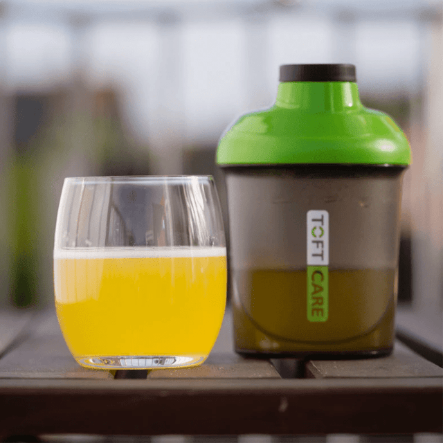 Shaker för proteinpulver med grön topp bredvid ett glas med gul dryck på ett träbord
