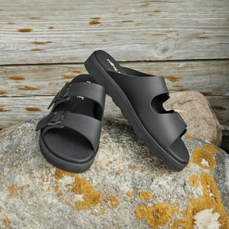 Två svarta New Feet sandaler på en mossig sten framför en trävägg, i en naturlig miljö