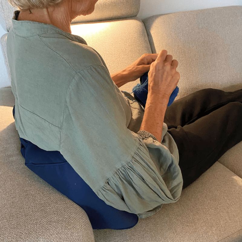 Äldre person sitter i soffa och stickar, med en blå ländkudde som stöd för ryggen
