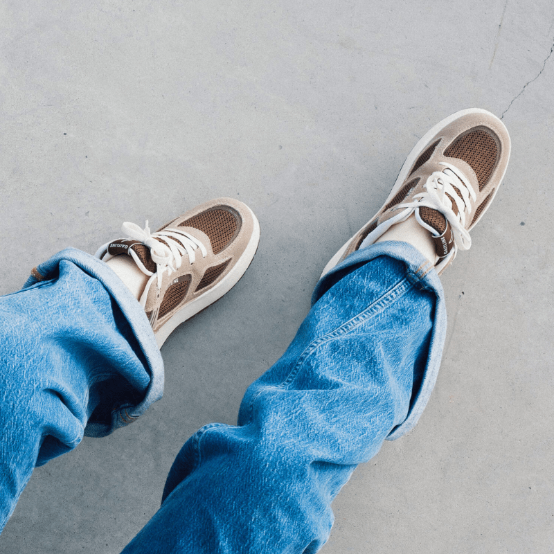 Fötter i bruna Gaitline Nova Lite damsneakers och upprullade blå jeans på en betongyta