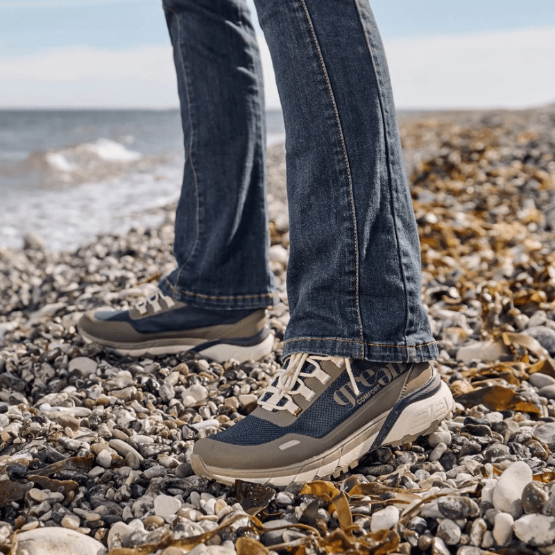 Person i blå jeans bär Green Comfort blå damsneakers på en stenstrand vid havet