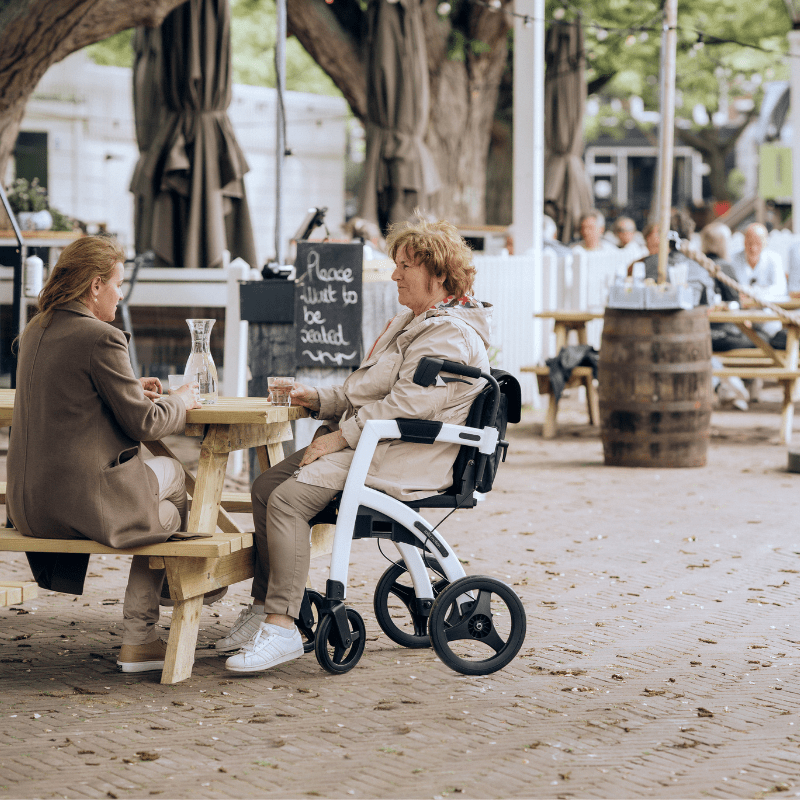 Äldre kvinna sitter vid ett utomhusbord med sin vita Rollz Motion rullstol, i samtal med en vän