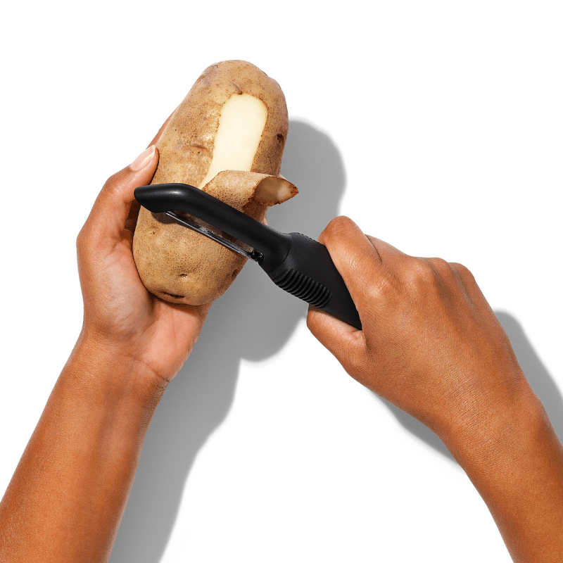 Händer skalar en potatis med en svart OXO potatisskalare, tydligt visar hur den används