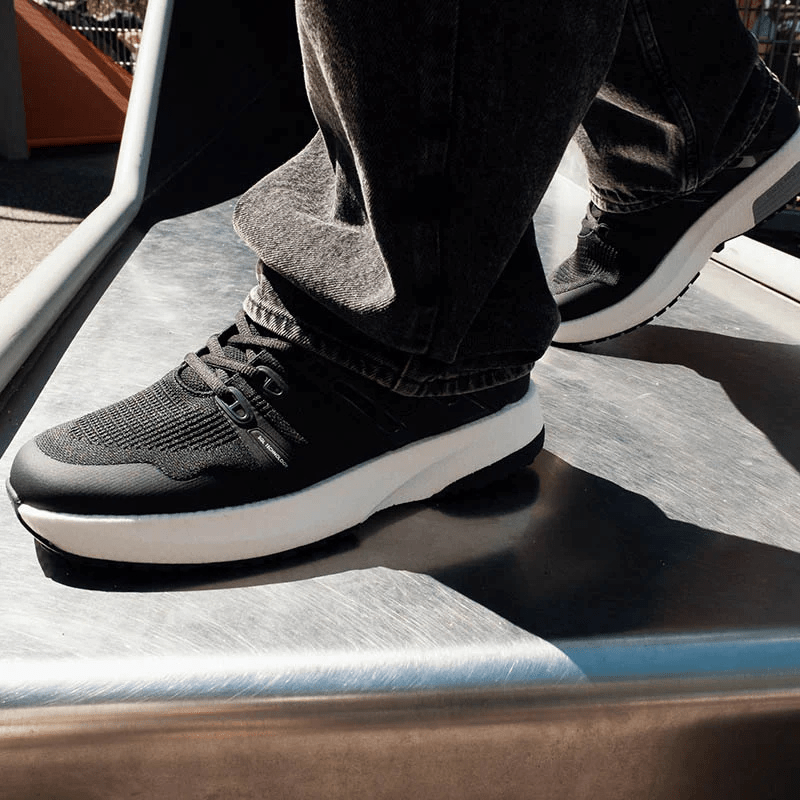 En person iført sorte Gaitline Nova Lite herresneakers går på et hårdt underlag