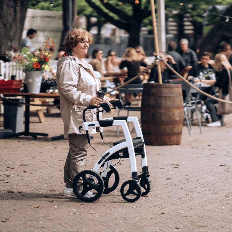 Äldre kvinna med en vit Rollz Motion rullator, stående utomhus på en kullerstensbelagd plats