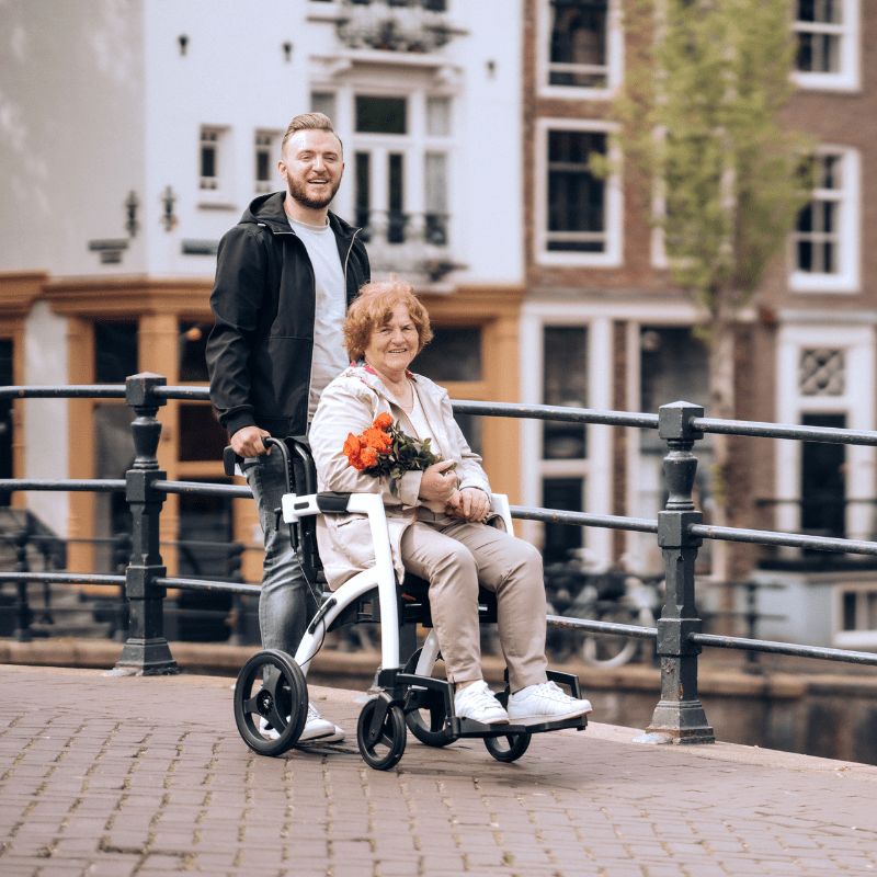 Man skjuter en äldre kvinna i en vit Rollz Motion rullstol över en bro, båda ler