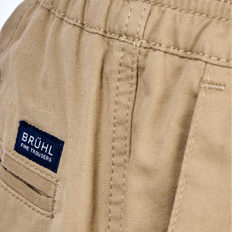 Närbild på resårmidjan och den blå Brühl "Fine Trousers"-etiketten på de beigefärgade byxorna