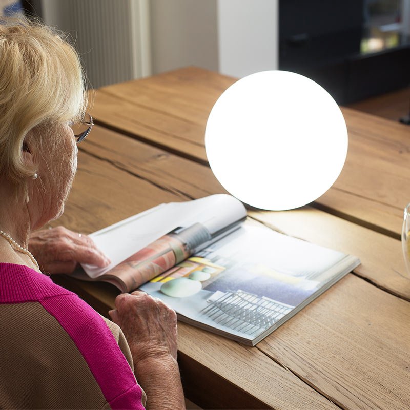 Senior kvinna läser tidning vid ett träbord med en tänd ljusterapilampa bredvid sig