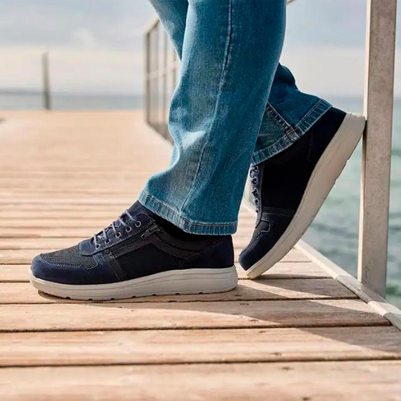 En person i blå jeans och mörkblå New Feet herrsneakers står på en träbrygga vid vattnet