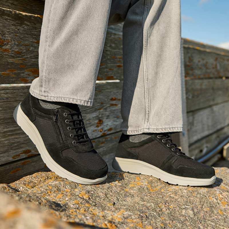 En person iført sorte New Feet herresko med grå bukser, stående på klipper udendørs