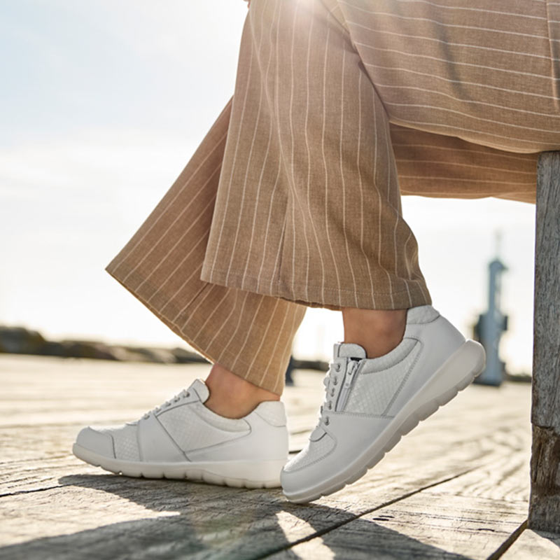 En person bär vita New Feet damsneakers med ränder på en träbrygga i solsken utomhus