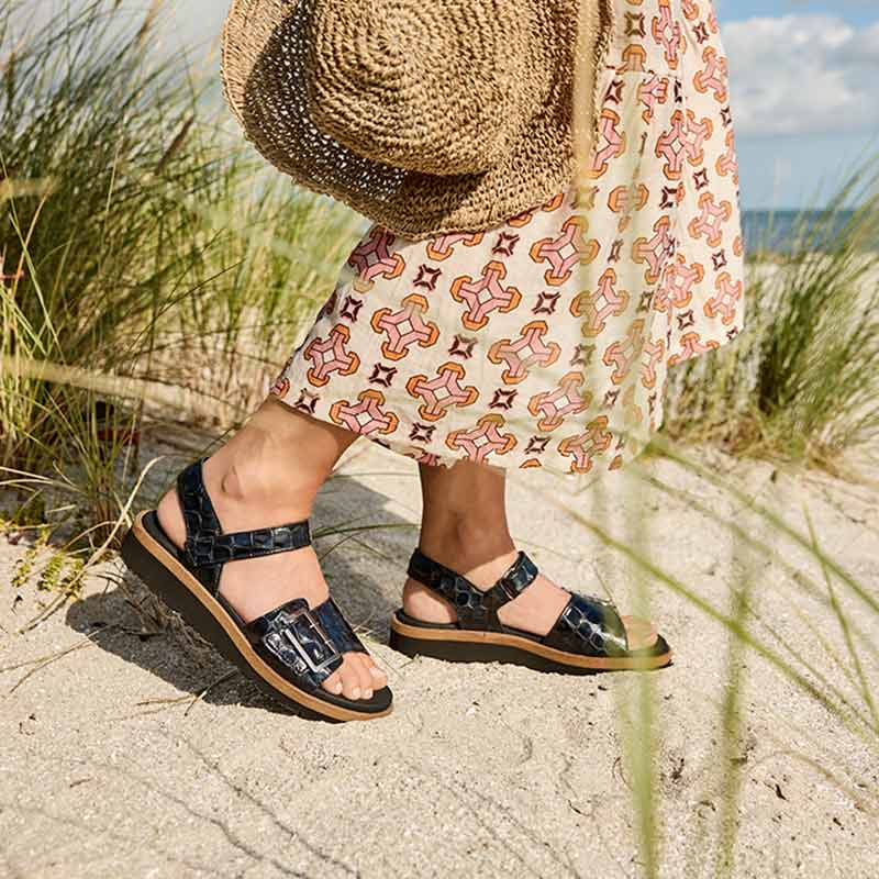 En kvinna bär svarta New Feet damsandaler med krokodilprägling på en sandstrand, med en stråväska