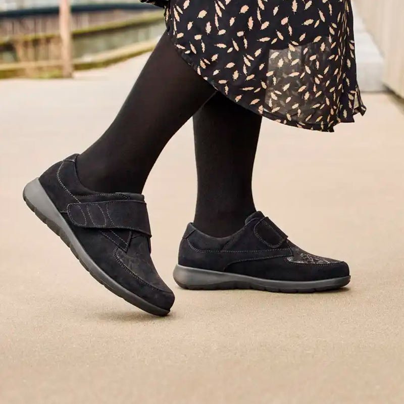En kvinna går utomhus med svarta New Feet damskor med kardborreband. Skorna har mönstrad ovansida