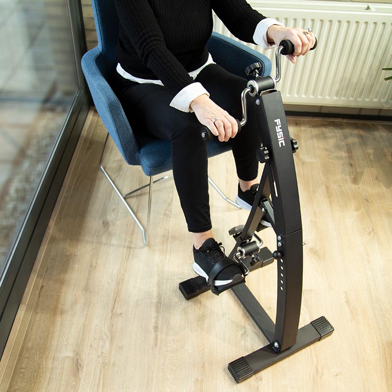 En person sitter i en stol och använder den svarta pedaltränaren för både arm- och benträning