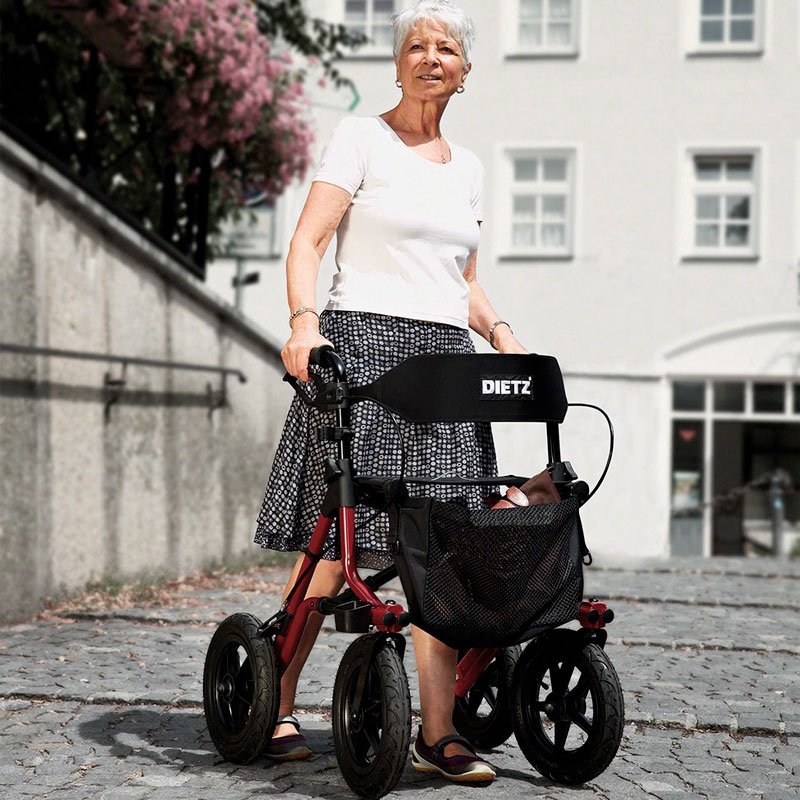 Äldre kvinna med grått hår använder en metallic-röd Taima XC utomhusrullator med Dietz ryggstöd på kullersten