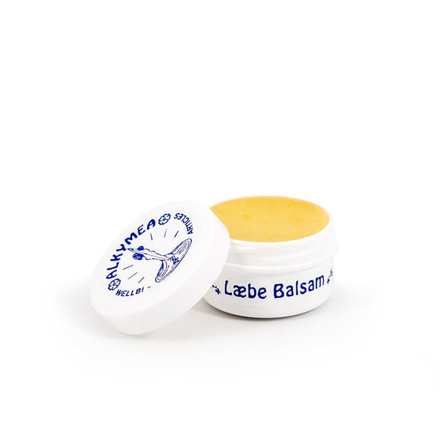Öppen vit burk med Alkymea vårdande läppbalsam som visar det gula balsamet inuti och den blå logotypen på locket