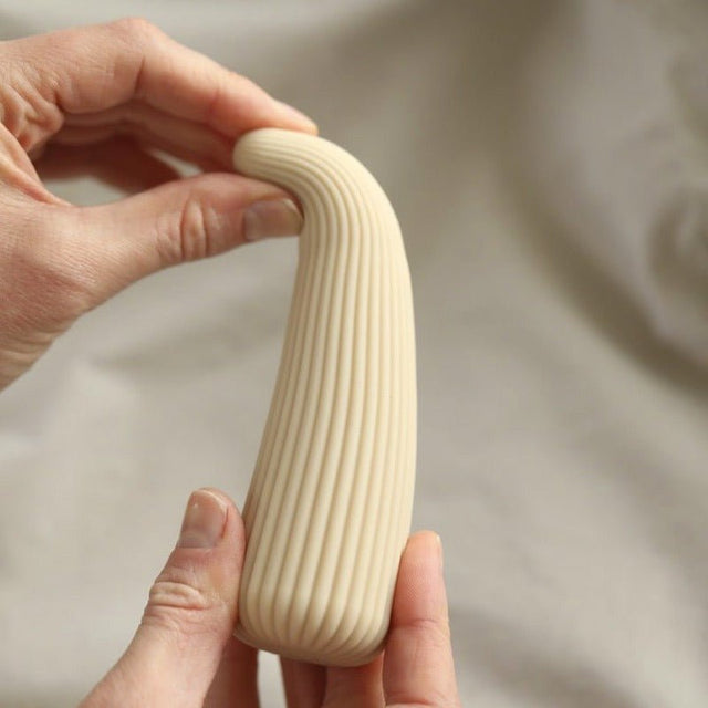 Händer som håller och böjer den beige Corny vibratorn, vilket visar dess flexibilitet och räfflade yta