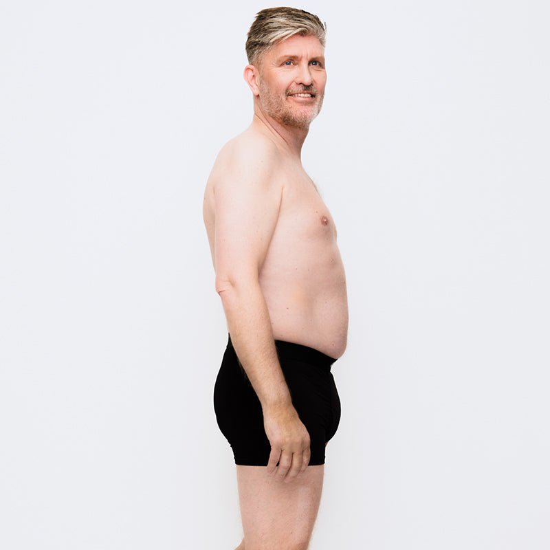 Medelålders man i svarta inkontinensboxershorts i mikrofiber, sedd från sidan mot vit bakgrund