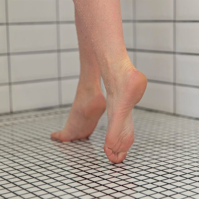 Bara fötter med vattendroppar på ett vitt klinkergolv i en dusch, som illustrerar halkskydd