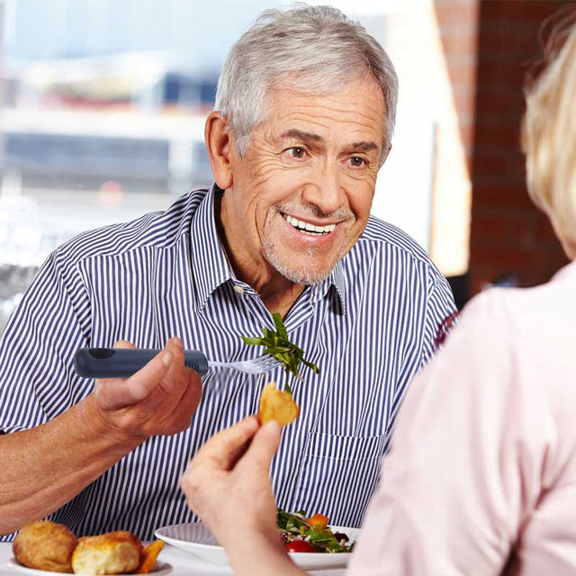 Äldre man med grått hår använder en gaffel med svart komfortgrepp för att äta sallad vid ett bord