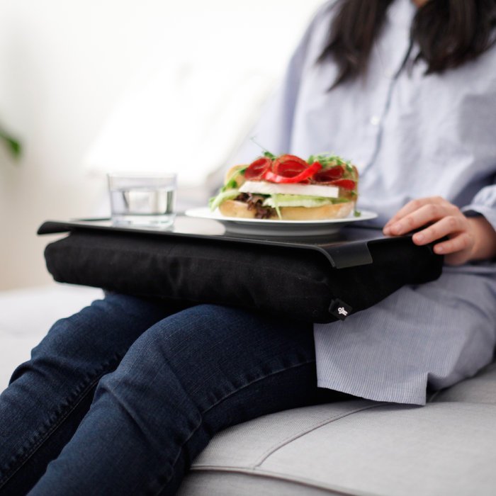 Person sitter i soffan med en svart bricka med kudde på knät, med en smörgås och ett glas vatten