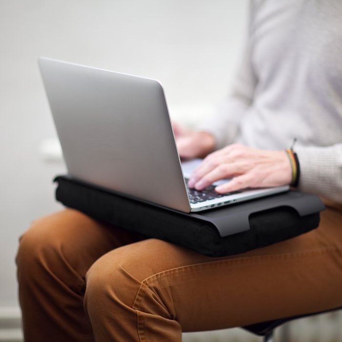 En person använder den svarta brickan med kudde som stabilt underlag för en silverfärgad laptop på knät