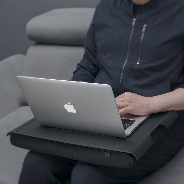 Person arbetar med en laptop på den svarta brickan med kudde i knät, sittande i en soffa