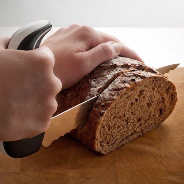 Händer skär en limpa mörkt bröd med den ergonomiska brödkniven med vinklat handtag på en skärbräda