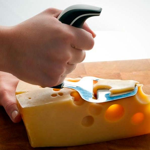 En hand använder den ergonomiska osthyveln för att skära en skiva gul ost med hål på en träskärbräda