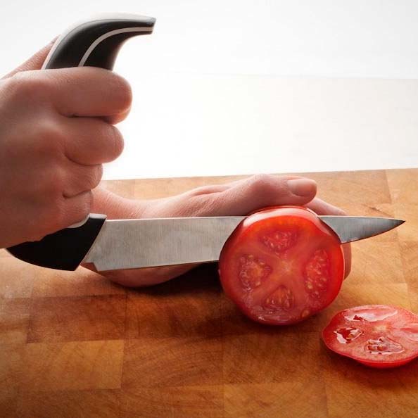 En hand använder den ergonomiska kökskniven för att skära en röd tomat på en träskärbräda