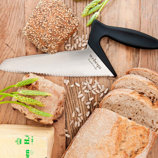 Ergonomisk brödkniv på ett träbord omgiven av skivat bröd, frallor och solrosfrön