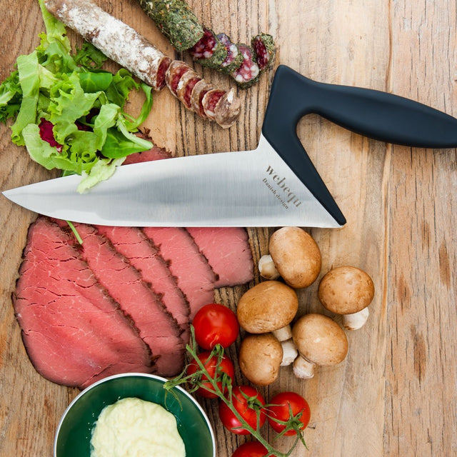 Ergonomisk kockkniv på en skärbräda med skivat kött, tomater och svamp, redo för matlagning