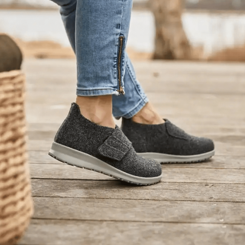 En person bär antracitgrå New Feet damtofflor i ull på ett trägolv utomhus