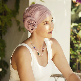 Kvinna med en ljusrosa Zuri turban i mjuk bomull med blomdetalj, sitter utomhus i vitt linne
